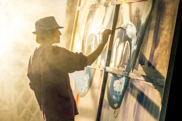 Cute middle aged womn graffiti artist dressed in work robe, jeans and hat. The woman artist is drawing the picture in street art style. She is holding paintbrush in hand. The female painter is standing sideways to the camera. Studio shooting in smoke