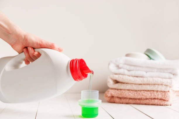 Close up of female hands pouring liquid laundry detergent into cap on white rustic table with towels on background in bathroom. stock photo