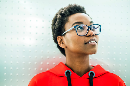 Young european african descent girl, in a red sweatshirt, looking up in front of a silver background. She wears glasses.