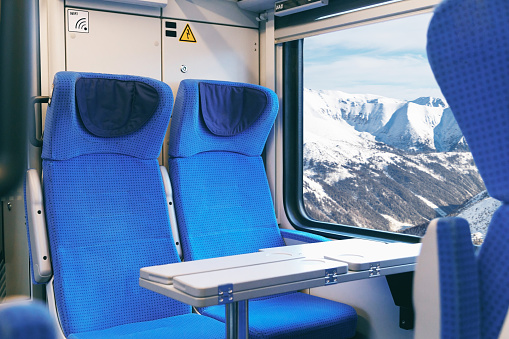Interior of an empty passenger train with blue chairs and mountain views outside the window while driving.