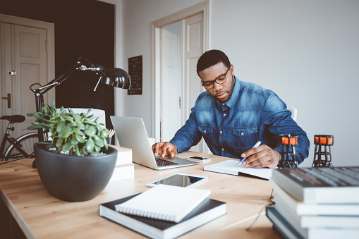 Shot of afro american young man in a home office using laptop and taking notes. Black guy sitting at table and working from home office.