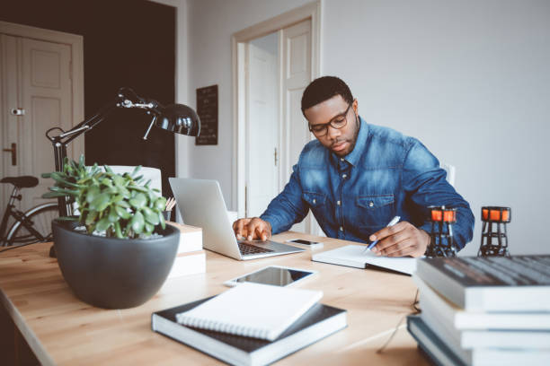 Afro american young man working at home office Shot of afro american young man in a home office using laptop and taking notes. Black guy sitting at table and working from home office. author stock pictures, royalty-free photos & images