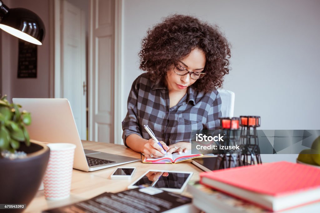 Afro american woman in a home office taking notes Shot of afro american woman in a home office taking notes in diary. Businesswoman sitting at table and working from home office. Writing - Activity Stock Photo