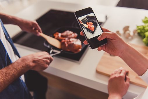 Beautiful young couple is cooking together in kitchen. Cropped image of girl taking photo of meal while her man is preparing it
