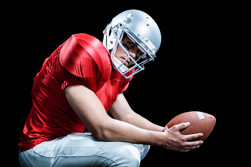 Portrait of American football player crouching while holding ball against black background