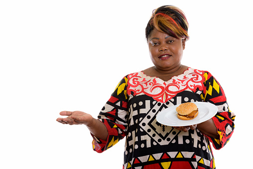 Studio shot of happy overweight black African woman smiling while showing something and holding burger served on white plate horizontal shot