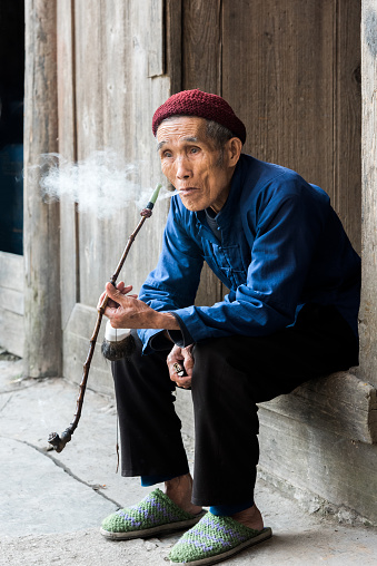 Very old chinese senior man sitting at the entrance door of his wooden home in a rural village, thinking while smoking a traditional handmade pipe. Real People Portrait. Zhao Xing, China