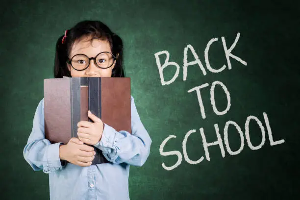 Cute schoolgirl looking at camera behind a book with back to school text on chalkboard