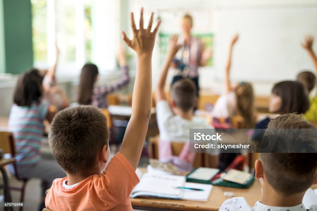 Back view of schoolboy raising hand to answer the question. Rear view of little boy and his classmates raising arms to answer teacher's question during the lecture in the classroom. Classroom Stock Photo