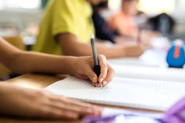 Close up of schoolgirl writing with pencil into notebook while learning in the classroom. There are people in the background.