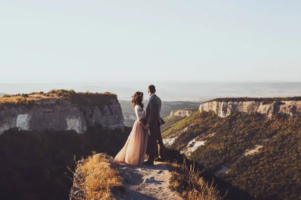 Photo of Bride and groom walking in mountains at sunset. Around the stunning scenery with views of the mountains and canyon Mangup