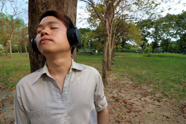 homem jovem feliz ouvindo música com fones de ouvido e encostado a uma árvore no parque público ao ar livre. - lying on side audio - fotografias e filmes do acervo