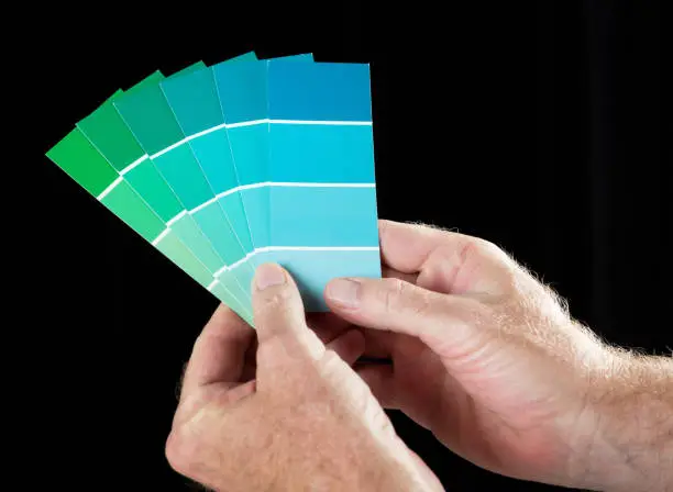 A man's hands hold up a fanned-out collection of paint colour samples in shades of blue and green against a black background with some copy space.