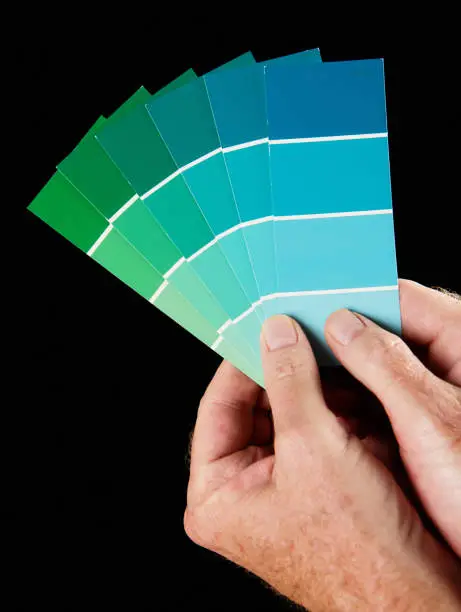 A man's hands hold up a fanned-out collection of paint colour samples in shades of blue and green against a black background.