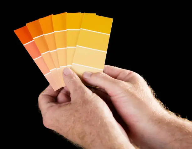 A man's hands hold up a fanned-out collection of paint colour samples in shades of yellow and orange against a black background with some copy space.