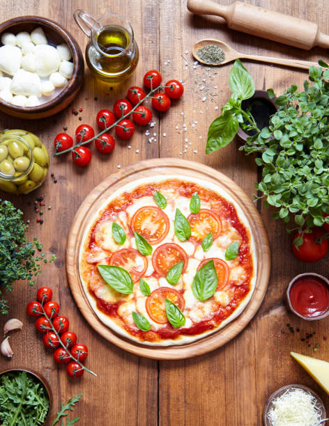 Home baked margarita pizza with fresh ingredients including tomatoes, mozzarella cheese, olives , basil and oil on a rustic wooden kitchen table. Italian food background. View from above.