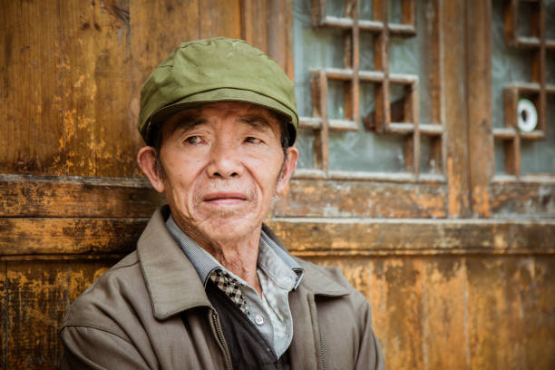 Chinese man in front of his home in Zhaoxing China Real People Portrait Chinese senior man sitting on bench in front of his home in a rural chinese village looking over. Chinese Real People Portrait. Zhaoxing, Guizhou, China mao original stock pictures, royalty-free photos & images