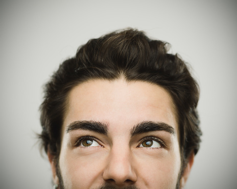 Close-up portrait of a real young confident man. Smiling male is against gray background and looking to the side. He has brown hair and beard. Horizontal studio photography from a DSLR camera. Sharp focus on eyes.
