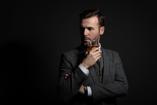 Studio portrait of successful man wearing tweed jacket, drinking whiskey, black background.