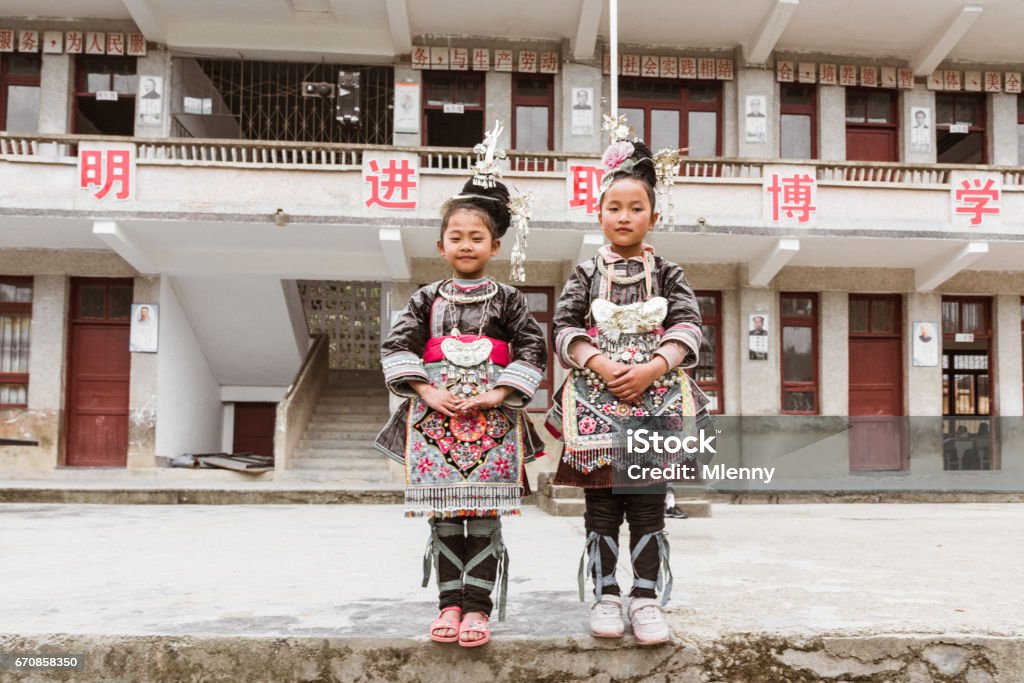 Chinese Dong People School Girls in Traditional Clothing Huanggang China Two chinese dong people school girls standing in traditional clothing together in front of their school building in the small Dong Village Huang Gang - Huanggang, Shuangjiang, Liping, Guizhou, China. Chinese Language Stock Photo