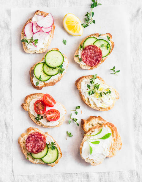 variety of mini sandwiches with cream cheese, vegetables and salami. sandwiches with cheese, cucumber, radish, tomatoes, salami, thyme, lemon zest on a light background, top view. flat lay - crostini imagens e fotografias de stock