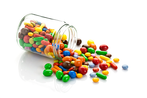 Open candy jar filled with multi colored chocolate covered peanut candies is lying down on white background. Some candies are spilled out of the jar directly on the background. DSRL studio photo taken with Canon EOS 5D Mk II and Canon EF 100mm f/2.8L Macro IS USM