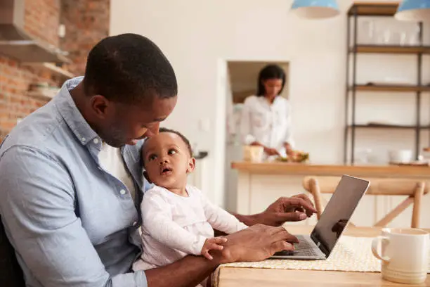 Photo of Father And Baby Daughter Use Laptop As Mother Prepares Meal
