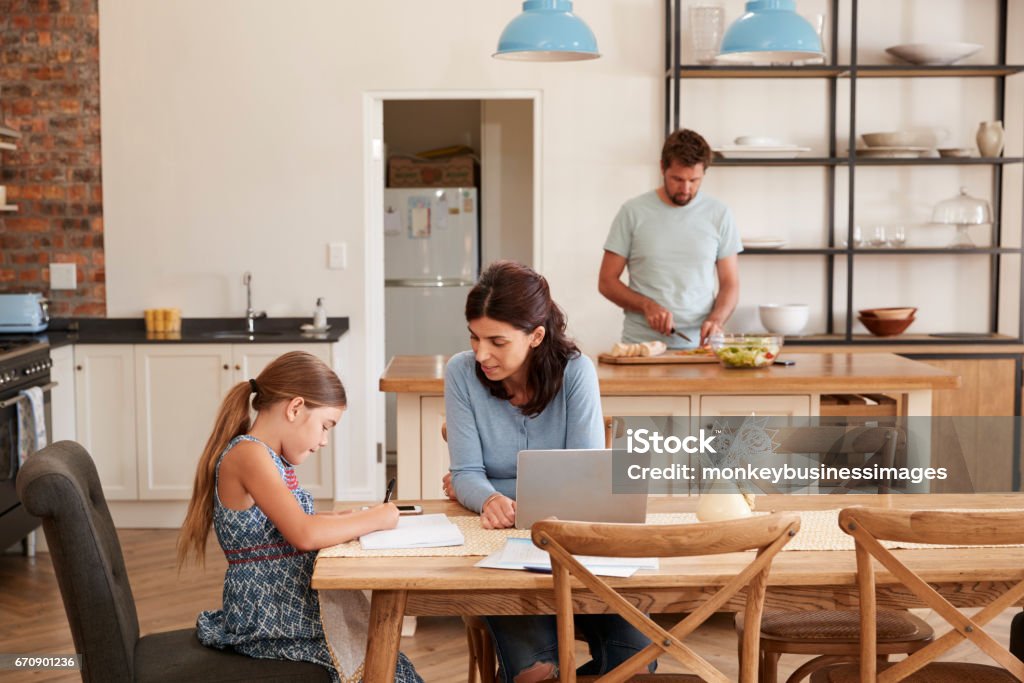 Mother Helps Daughter With Homework As Father Makes Meal Family Stock Photo