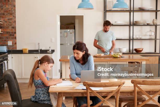 Mother Helps Daughter With Homework As Father Makes Meal Stock Photo - Download Image Now