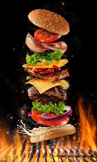 Flying burger ingredients above grill fire. Concept of low gravity motion and meal preparation. Isolated on black background