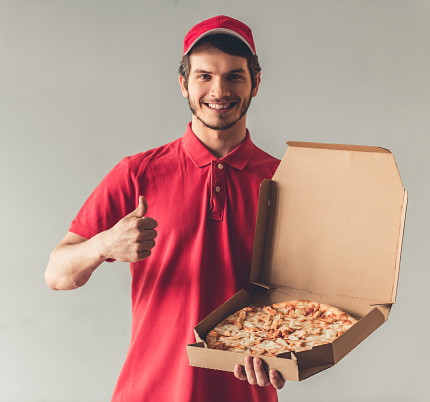 Handsome young delivery worker in red uniform is holding a pizza, looking at camera and smiling, on gray background