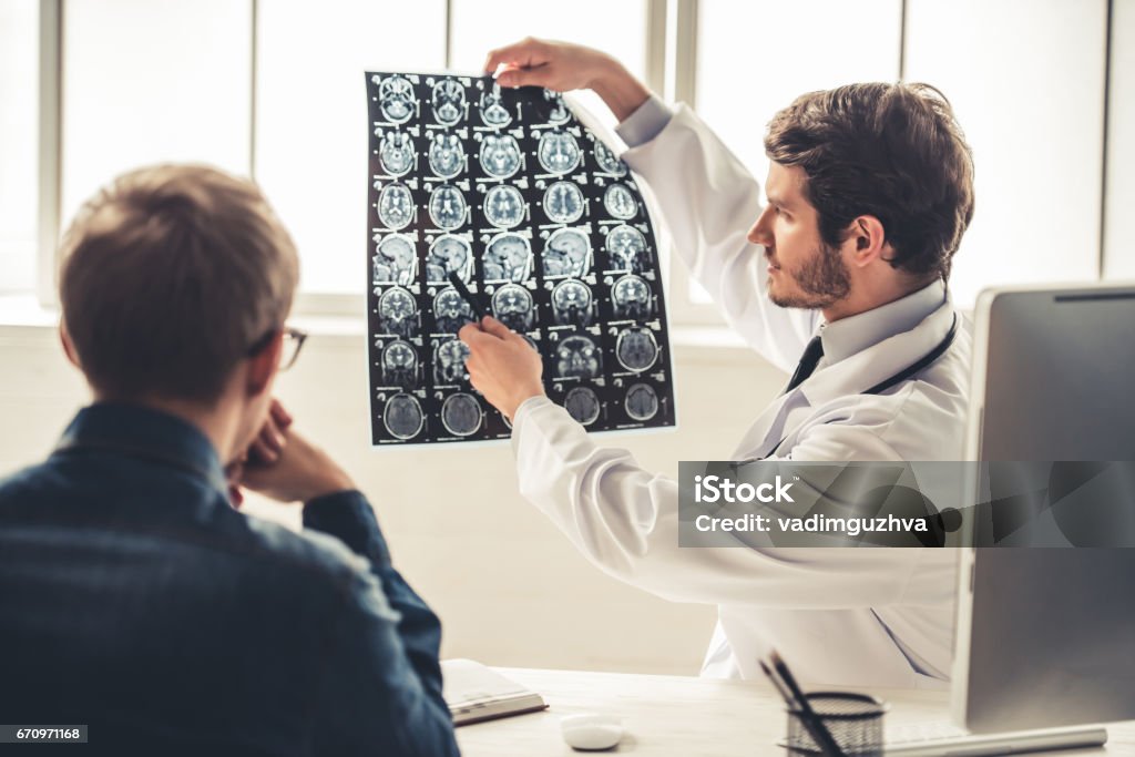 At the doctor Handsome young doctor in white coat is showing x-ray image to his patient while working in office Patient Stock Photo