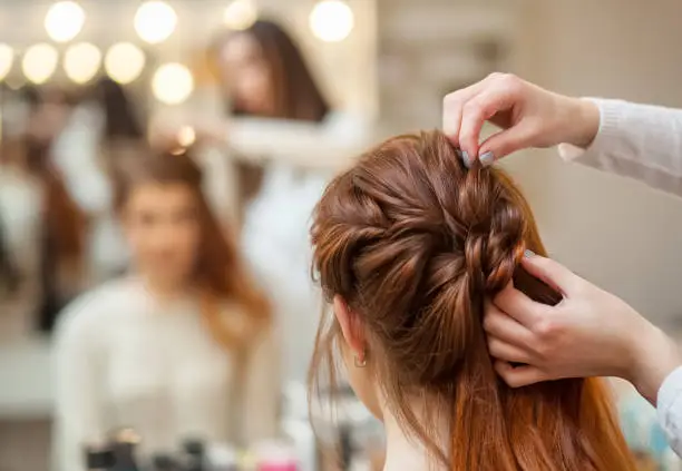 Photo of Beautiful, red-haired girl with long hair, hairdresser weaves a French braid, in a beauty salon