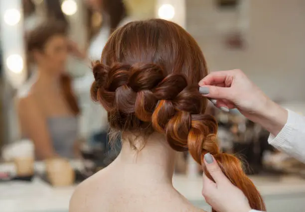 Beautiful, with long, red-haired hairy girl, hairdresser weaves a French braid, close-up in a beauty salon. Professional hair care and creating hairstyles.