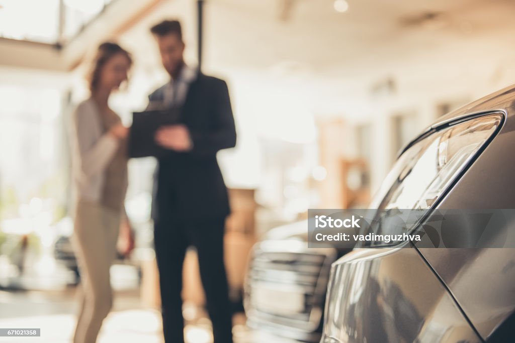 Visiting car dealership Beautiful young woman is talking to handsome bearded sales manager while choosing a car in dealership Car Stock Photo