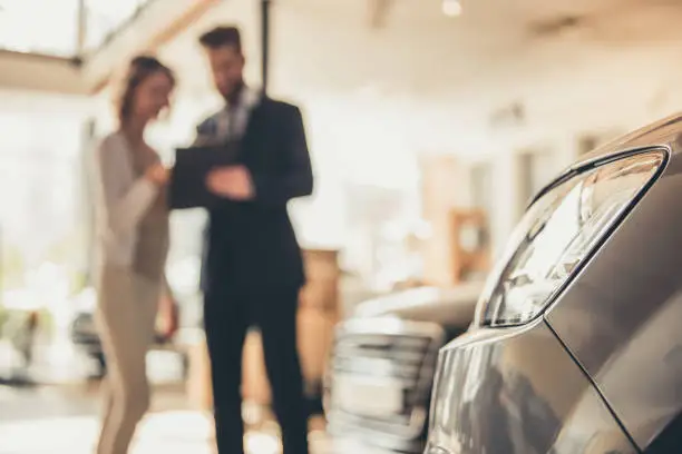 Beautiful young woman is talking to handsome bearded sales manager while choosing a car in dealership