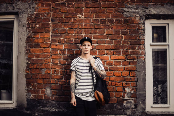 れんが造りの壁の横にキャップで魅力的な男 - tattoo t shirt brick wall portrait ストックフォトと画像