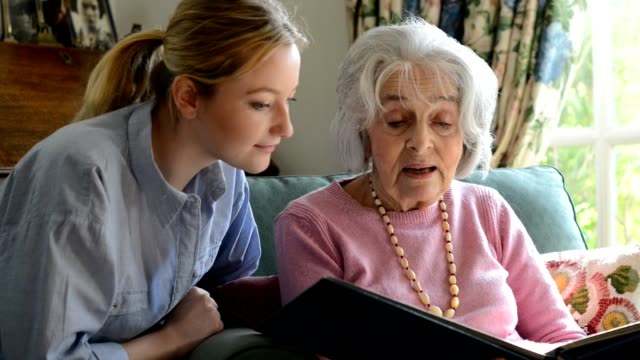 Senior woman sitting with adult granddaughter at home looking through photo album together