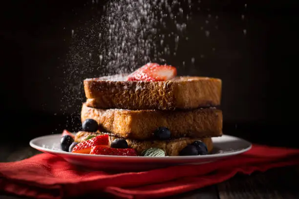 French toast with fresh strawberries, blueberries,mint, syrup and powdered sugar being sprinkled over the breakfast.