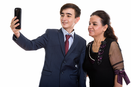 Studio shot of happy young Persian teenage businessman and mature Persian woman smiling while taking selfie picture with mobile phone together horizontal shot