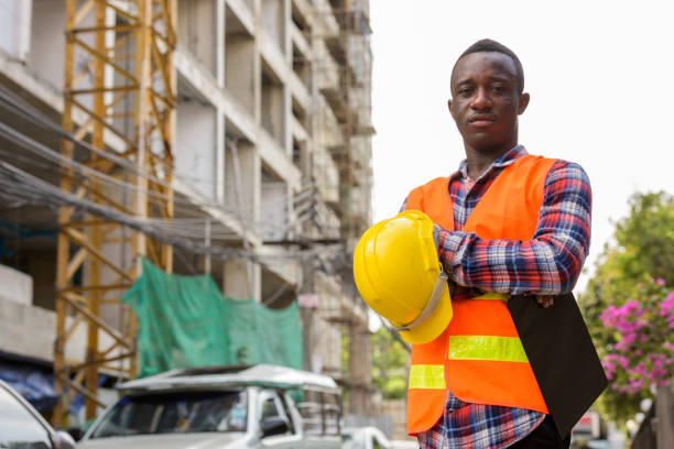 istock Young black African man construction worker holding clipboard and hard hat with arms crossed at building site 671124094