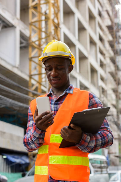 istock Young happy black African man construction worker smiling while holding clipboard and using mobile phone at building site 671124240