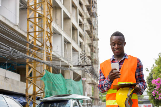 istock Young happy black African man construction worker smiling and using mobile phone while holding safety helmet and clipboard at building site 671124324