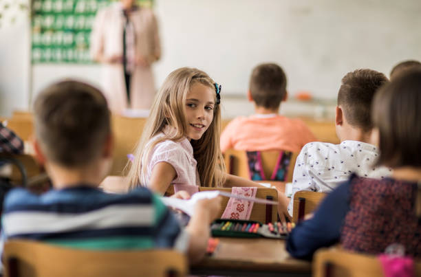 istock Smiling little girl taking paper from classmate in the classroom. 671176366
