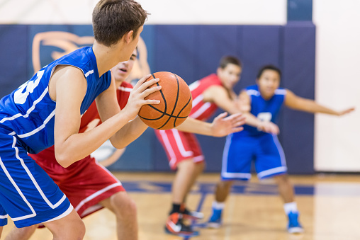 A junior basketball player is standing on basketball court with a ball during the training and looking at the camera. Portrait of a young athlete with a basketball on court. In background is his team.
