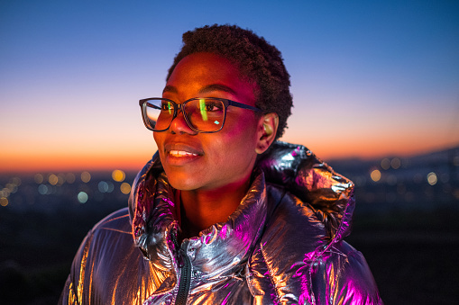Young girl outdoors wearing eyeglasses and a shiny jacket that reflects colorful lights. Photo taken at disk, with city lights defocused at background.