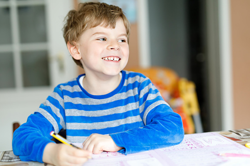 Portrait of cute happy school kid boy at home making homework. Little child writing with colorful pencils, indoors. Elementary school and education. Kid learning writing letters and numbers.