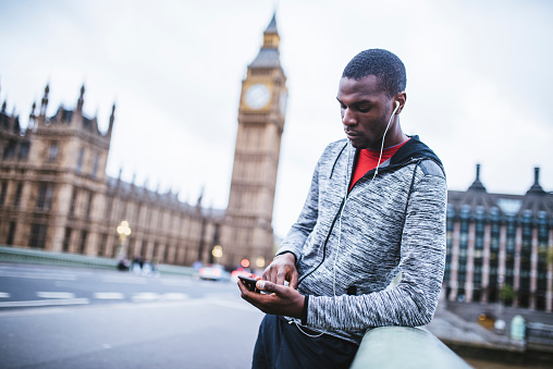 African-American man standing outdoors in the city. He is focused and determined. He is using his mobile phone. Big ben in the back.