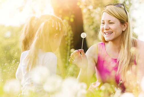 Smiling young mother is holding a dandelion flower for her little daughter while sitting in meadow. Real family, genuine emotions. Surface level shot.