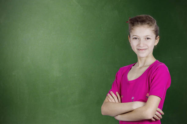 Closeup portrait thinking confident young, happy, smiling  student girl, woman by green chalkboard background, arms crossed, high school pupil, contemplating looking at you. Positive face expressions